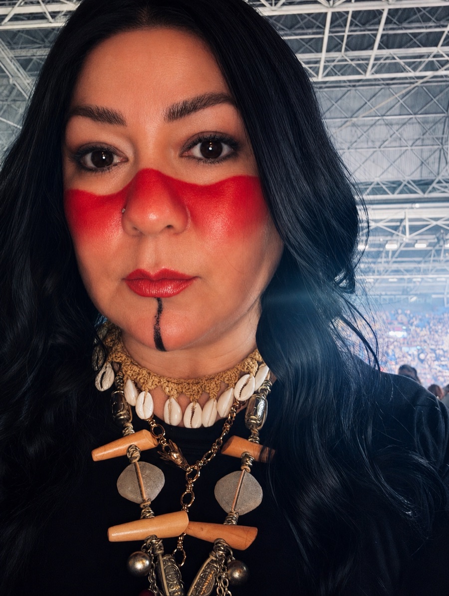 Close-up portrait with red face paint, dark hair, and layered shell and metal jewelry.